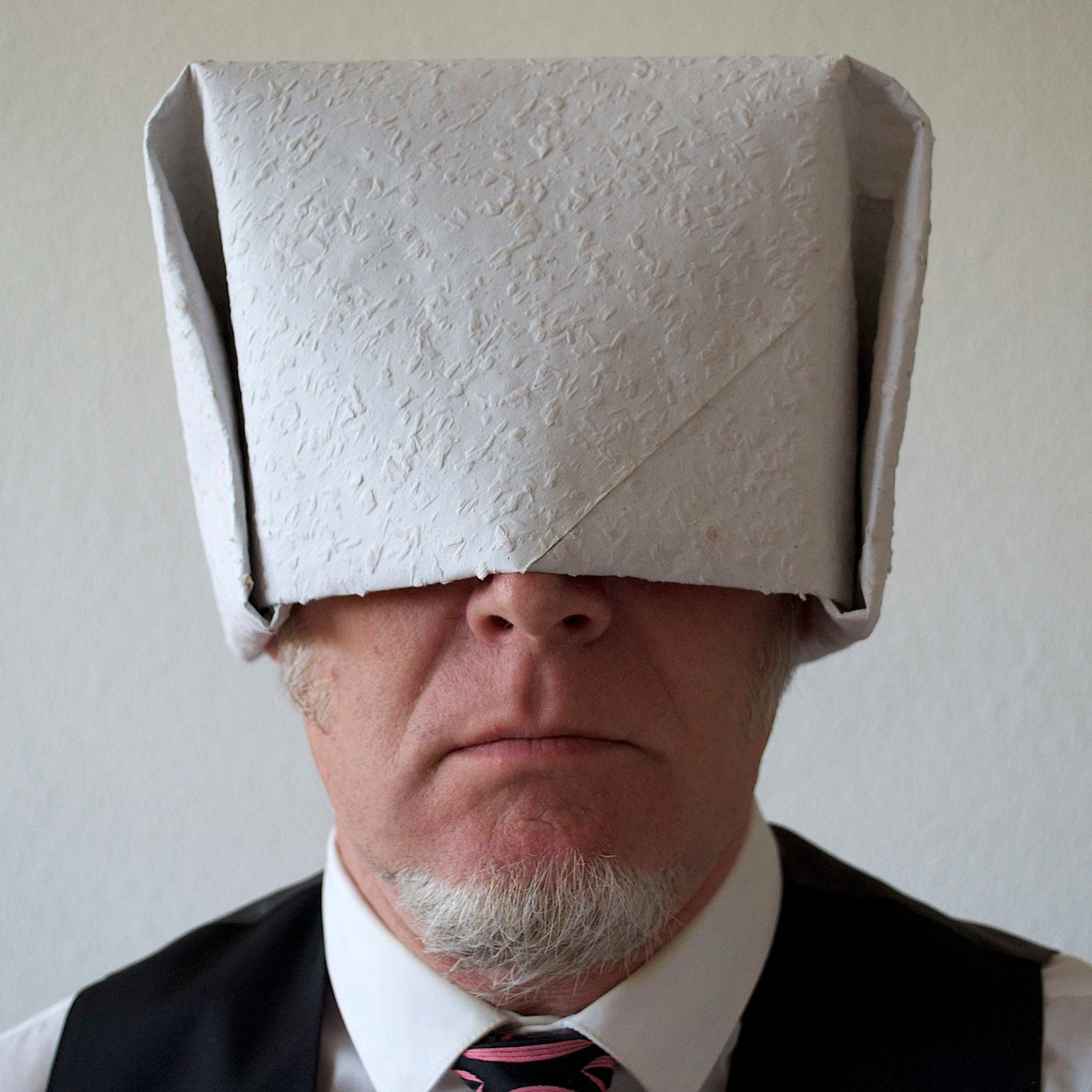Portrait von Frank Bölter. Frank Bölter trägt als Kopfbedeckung einen Hut aus Raufasertapete, der tief ins Gesicht gezogen ist, so dass man das Gesicht nur noch von der Nase an abwärts sehen kann. Bekleidet ist Herr Bölter mit einem weißen Hemd, einer schwarzen Weste und einer Krawatte. Herr Bölter ist Bartträger.