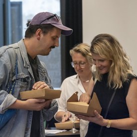 Drei Gäst*innen beim Öffnen einer Schachtel aus brauner Pappe. Die Schachteln stammt von der Food Performance und enthalten jeweils einen Snack.