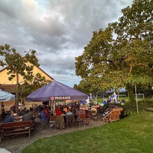 Biergarten mit vielen Menschen an Holztischen unter großen Sonnenschirmen. Im Hintergrund, auf einer kleinen Wiese neben Bäumen und Spielgeräten, steht eine beleuchtete Bühne mit Musikern.