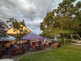 Biergarten mit vielen Menschen an Holztischen unter großen Sonnenschirmen. Im Hintergrund, auf einer kleinen Wiese neben Bäumen und Spielgeräten, steht eine beleuchtete Bühne mit Musikern.