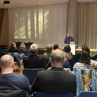 Vortrag in einem Saal: Menschen sitzen in blauen Stühlen, hören dem Impulsgeber Jacob Sylvester Bilabel am Pult zu. Auf der Leinwand ein Waldbild, daneben ein Kultur-Banner.
