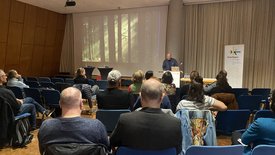 Vortrag in einem Saal: Menschen sitzen in blauen Stühlen, hören dem Impulsgeber Jacob Sylvester Bilabel am Pult zu. Auf der Leinwand ein Waldbild, daneben ein Kultur-Banner.