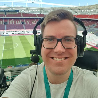 Mann mit Brille und Headset lächelt in die Kamera, im Hintergrund ein Fußballstadion mit grünem Spielfeld und leeren Tribünen.