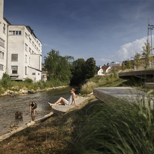 Foto: Blick auf die terassenförmig angelegten Sitz- und Liegemöglichkeiten an der Fils. Auf der anderen Seite der Fils ist ein großes Gebaüde zu sehen. Ein Kind Spielt im Wasser, eine Frau, vermutlich die Mutter, schaut vom Ufer aus zu