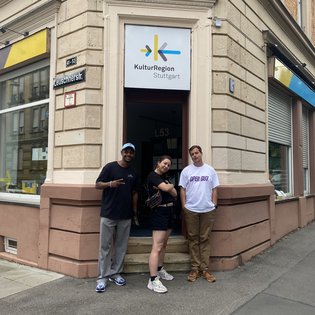 B-Girl Sophie Manuela Lindner, Popping Tänzer Milyon Hagos und Moderator Ingmar Volkmann vor der Geschäftsstelle der KulturRegion Stuttgart. Über der Tür ist das Logo der KulturRegion zu sehen.
