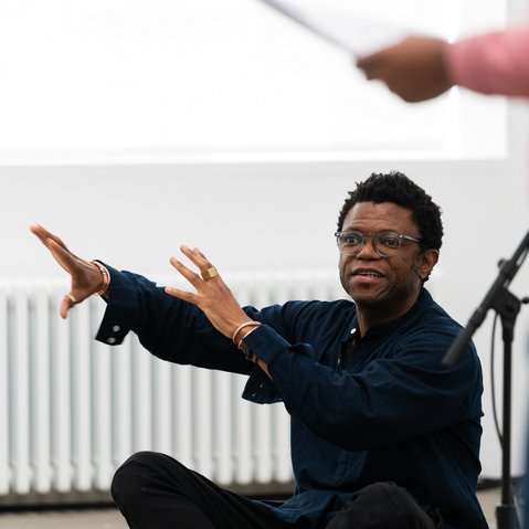 Neo Muyanga, ganz in Schwarz gekleidet, sitzt im Schneidersitz auf dem Boden und richtet große Gesten an eine oder mehrer Personen, die scheinbar mit ihm im Raum sind.