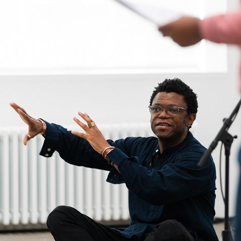 Neo Muyanga, ganz in Schwarz gekleidet, sitzt im Schneidersitz auf dem Boden und richtet große Gesten an eine oder mehrer Personen, die scheinbar mit ihm im Raum sind.