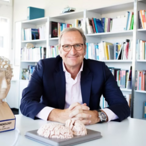 Dr. h.c. Lutz Jäncke sitzt an einem weißen Tisch und hat die Hände auf dem Tisch gefaltet. Er trägt eine Brille und ist mit einem weißen Hemd und ein blauen Jacket bekleidet. Vor ihm und neben ihm auf dem Tische stehen Modelle des Gehirns. Im Hintergrund ist ein großes Bücherregal zu sehen.