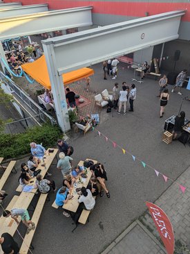 Blick von oben auf ein Fest. Mehrere Personen sitzen an Bierbängen und -tischen. Bunte Fahnengirlanden sind über dem fest gespannt. 