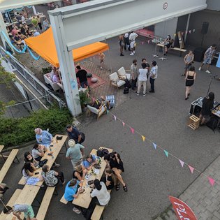 Blick von oben auf ein Fest. Mehrere Personen sitzen an Bierbängen und -tischen. Bunte Fahnengirlanden sind über dem fest gespannt. 