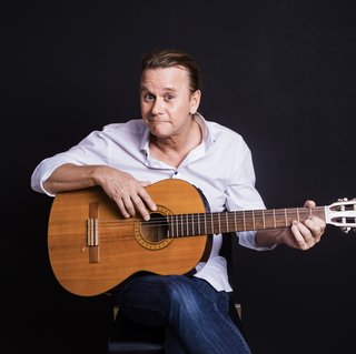 Nicolai Köppel sitzt mit zurückgebundenen Haaren vor einem dunklen Hintergrund und hält eine klassische Akustikgitarre. Er trägt ein weißes Hemd und Jeans, schaut direkt in die Kamera und lächelt leicht. Seine linke Hand greift in die Saiten, während die rechte Hand auf dem Korpus der Gitarre ruht.