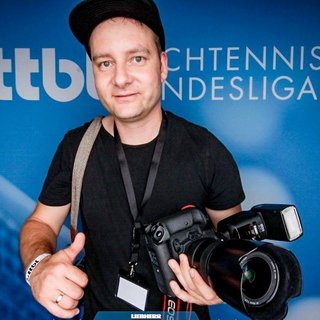 Person mit schwarzem T-Shirt hält eine professionelle Kamera mit Blitz vor einem blauen Hintergrund mit dem Schriftzug 'ttbl' und 'TISCHTENNIS BUNDESLIGA'