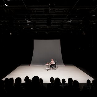 Blick auf die komplette Bühne von »Precarious Moves«. In der Mitte der Bühne Michael Turinsky im Rollstuhl, der einen Servierwagen mit Getränken darauf vor sich herschiebt.
