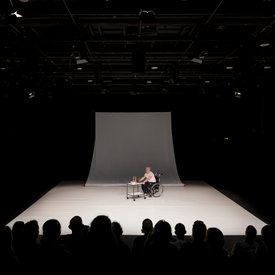 Blick auf die komplette Bühne von »Precarious Moves«. In der Mitte der Bühne Michael Turinsky im Rollstuhl, der einen Servierwagen mit Getränken darauf vor sich herschiebt.