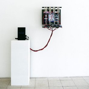 Eine schwarze PlayStation 2 steht auf einem weißen Sockel vor einer weißen Wand. Ein dicker Kabelstrang verbindet die Konsole mit einer mechanischen Vorrichtung, die an der Wand montiert ist. In der Vorrichtung befindet sich ein Gamecontroller, auf dessen Tasten mehrere kleine Motoren oder Aktuatoren montiert sind. Die Kabel und mechanischen Bauteile wirken industriell und technisch. Der Raum ist minimalistisch gestaltet mit einem hellen Fliesenboden.