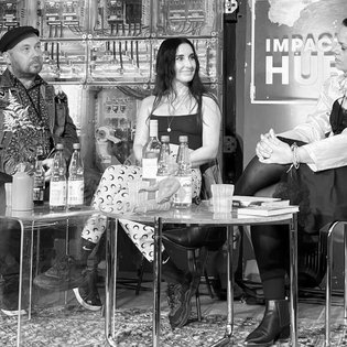 Mirna Funk sitzt mit überschlagenen Beinen zwischen Yuriy Gurzghy und Anna Lampert auf der Bühne. Sie lächelt Moderatorin Anna Lampert zu, die gerade das Wort an sie zu richten scheint.