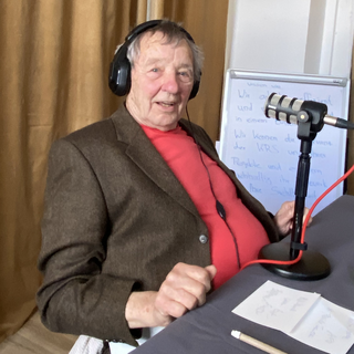 Gast Peter Grohmann während der Aufnahme des Podcast. Er sitzt an einem Tisch, trägt Kopfhörer und hat ein Tischmikrofon vor sich stehen. Peter Grohmann trägt ein rotes Shirt und ein braunes Jacket.