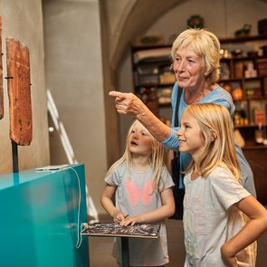 Blick in die Ausstellung. Eine ältere Dame betrachtet mit zwei Mädchen ein Ausstellungsobjekt.