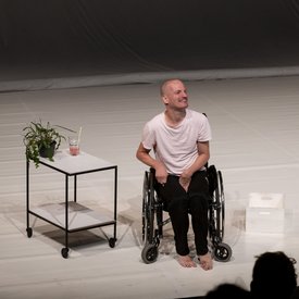 Michael Turinsky im Rollstuhl auf der Bühne. Neben ihm steht ein Servierwagen mit einem Getränk.