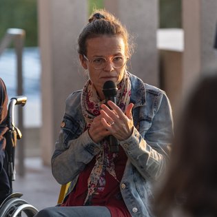 Anke Hofmann auf der Terrasse des Literaturmuseum der Moderne. Sie hat ihre Haare zu einem Knoten zusammengebunden und trägt eine Brille. Bekleidet ist sie mit einer Jeansjacke und einem bunten Seidenschal. Sie spricht in ein Mikrofon.