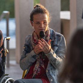 Anke Hofmann auf der Terrasse des Literaturmuseum der Moderne. Sie hat ihre Haare zu einem Knoten zusammengebunden und trägt eine Brille. Bekleidet ist sie mit einer Jeansjacke und einem bunten Seidenschal. Sie spricht in ein Mikrofon.