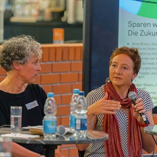 Dr. Birte Werner und Jule Fiedler auf dem Podium,im Hintergrund zeigt ein Bildschirm den Titel: Sparen wir uns kaputt?