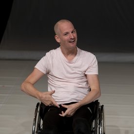 Michael Turinsky auf der Bühne. Er nutzt einen Rollstuhl und trägt ein helles Shirt und eine schwarze Hose.
