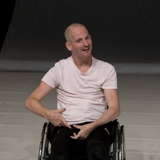 Michael Turinsky auf der Bühne. Er nutzt einen Rollstuhl und trägt ein helles Shirt und eine schwarze Hose.