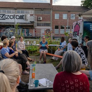 Gruppe von Menschen sitzt in einem Kreis im Freien, offenbar in einem Garten- oder Gemeinschaftsraum. Auf den Tischen stehen Getränke, Becher und Papiere. Im Hintergrund sind ein Graffiti-verziertes Gebäude mit dem Schriftzug ‚zollamt‘ sowie ein farbenfrohes Wandbild auf einer Struktur zu sehen – die Szene vermittelt eine offene, gemeinschaftliche Atmosphäre.