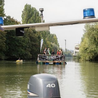 Das Festival-Floß auf einem Gewässer. Fotografiert vom Begleitboot der DLRG aus, weshalb hiervon noch der Motor und Blaulichter zu sehen sind.