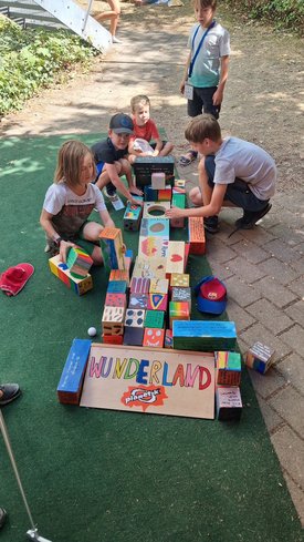 Fünf Kinder spielen draußen mit bunten Holzklötzen, die zu einer langen Reihe mit dem Schild 'WUNDERLAND' vorne aufgebaut sind