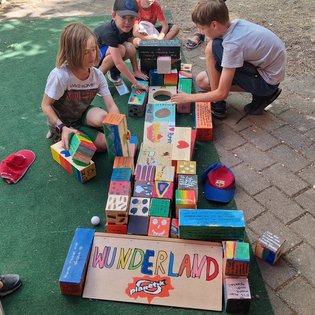 Fünf Kinder spielen draußen mit bunten Holzklötzen, die zu einer langen Reihe mit dem Schild 'WUNDERLAND' vorne aufgebaut sind