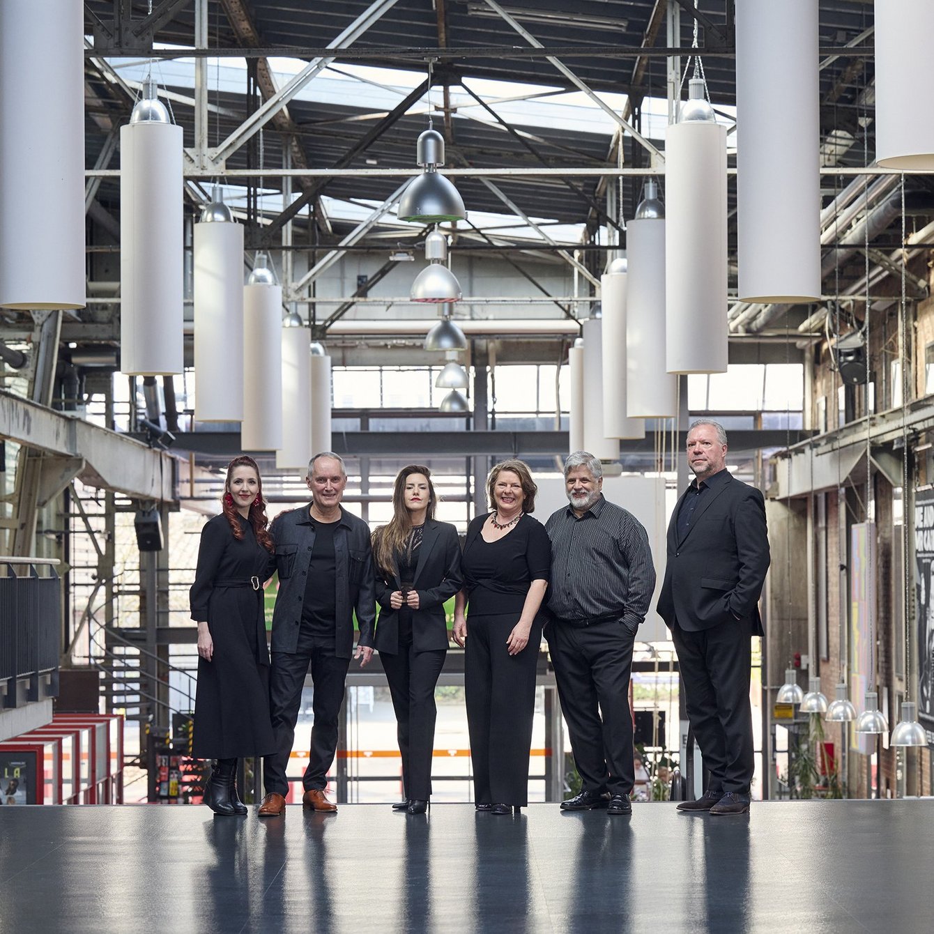 Sechs Personen in schwarzer Kleidung stehen in einer hellen Industriehalle mit hohen Decken und großen Fenstern nebeneinander