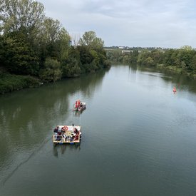 Das Festival-Floß unterwegs auf einem Gewässer in der Region. Gezogen wird es von einem motorisierten Begleitboot. An den Ufern des Gewässers stehen Bäume und Büsche. Das Bild wurde anscheinend von einer Brücke aus aufgenommen.