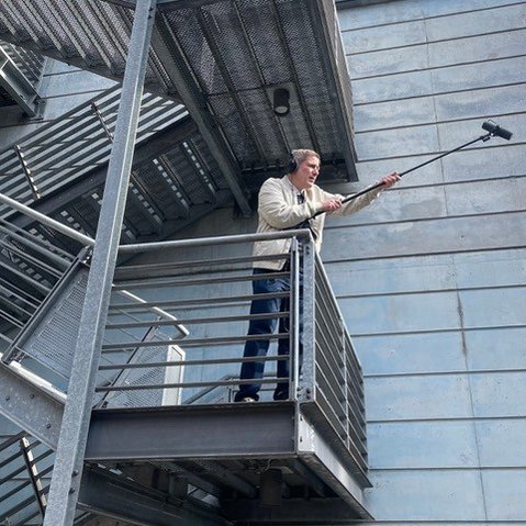 Person steht auf einem Metallbalkon vor einer grauen Wand und hält eine lange Stange mit einem Mikrofon nach vorne.