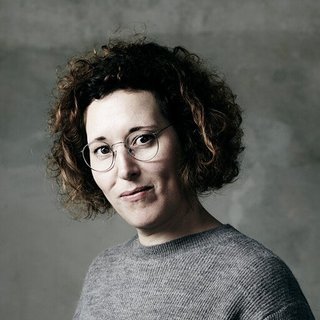 Person mit lockigem Haar trägt einen grauen Pullover und schwarze Hose und steht vor einer grauen Wand mit einer Hand in der Hosentasche