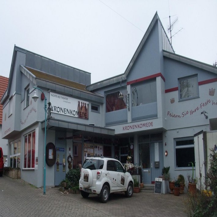 Außenansicht des Gebäudes „KronenKomede“ in Filderstadt, Deutschland. Es handelt sich um ein Theaterhaus mit der Aufschrift „NICHTS ALS THEATER“ und „Feiern Sie Ihre Feste hier“. Ein weißes Auto mit dem Logo des Theaters steht vor dem Eingang. Die Fassade ist hellgrau mit roten Akzenten, dekoriert mit Theatermotiven und Pflanzen.