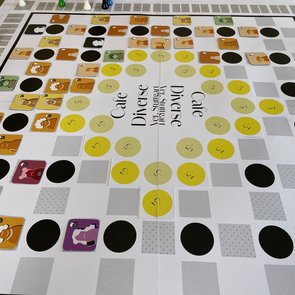 Ein buntes Brettspiel mit dem Titel „Café Diverse“ ist auf einem Tisch aufgebaut. Das Spielfeld zeigt viele farbige Kreise und Quadrate in Gelb-, Grau- und Schwarztönen, auf denen kleine quadratische Plättchen mit Comicfiguren liegen. Um das Spielfeld herum stehen Spielfiguren in verschiedenen Farben (Gelb, Grün, Weiß). Am Rand befindet sich eine Zählleiste mit Zahlen. In der Mitte des Spielfelds steht mehrfach der Schriftzug „Café Diverse – Yes, Sürgent Yes! Stuttgart“.