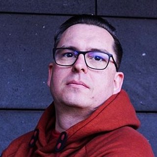  Eine Person mit kurz geschnittenem, dunkelbraunem Haar steht vor einer Wand aus dunkelgrauen, rechteckigen Fliesen. Die Person trägt eine rote Kapuzenjacke und eine Brille mit dunklem Rahmen. Sie blickt direkt in die Kamera mit neutralem Gesichtsausdruck. Die Beleuchtung ist gleichmäßig, und es sind keine weiteren Objekte im Hintergrund sichtbar.