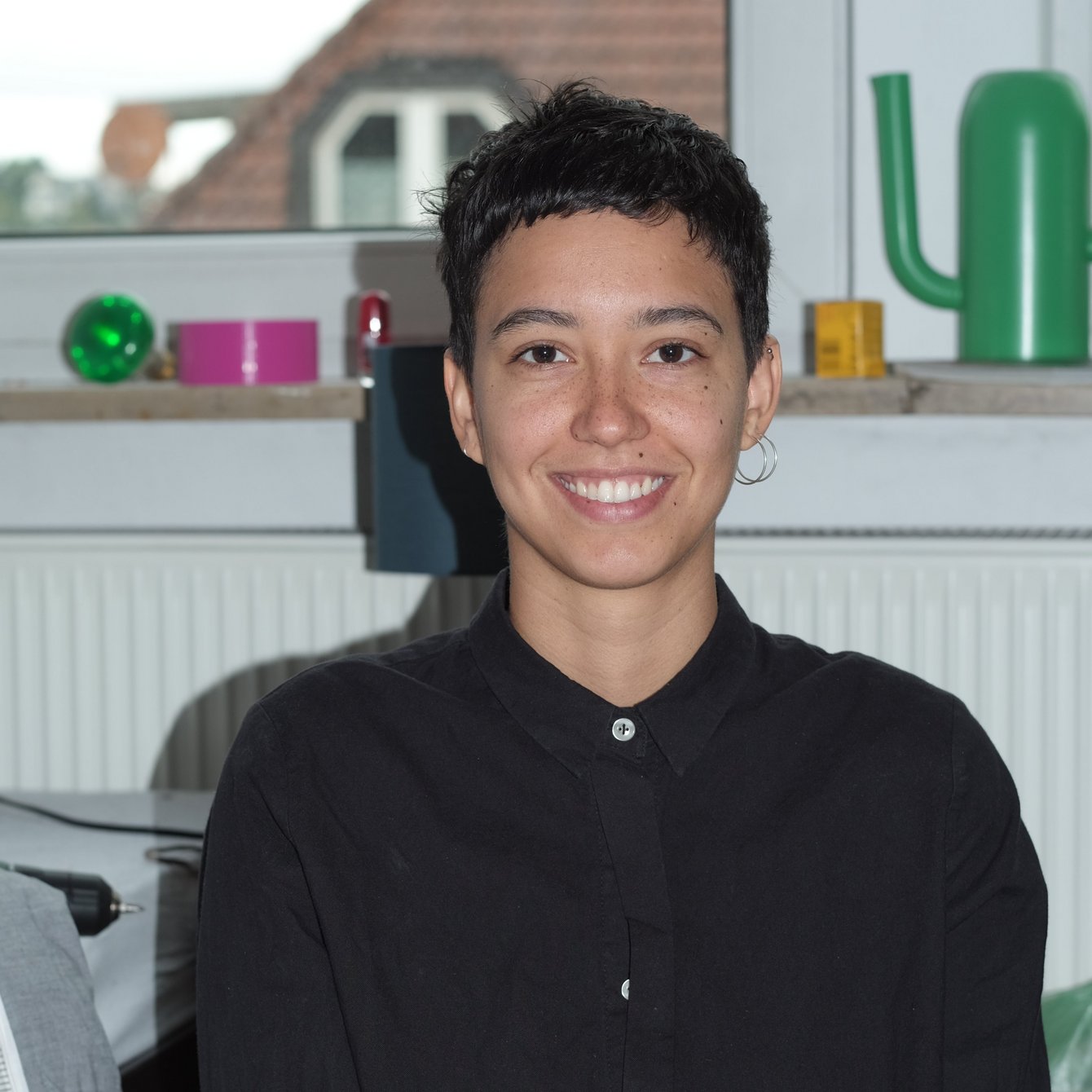 Junge Person mit kurzen dunklen Haaren und schwarzem Hemd sitzt lächelnd vor einem Fenster mit Dekoobjekten