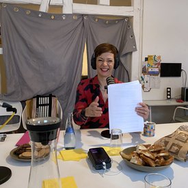 Moderatorin Kimsy von Reischach während der Podcast-Aufnahme. Sie sitzt an einem Tisch, lacht fröhlich in die Kamera, trägt Kopfhörer und hält Papierunterlagen in der Hand.
