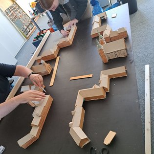 Mehrere Personen bauen auf einem Tisch ein Modell aus Pappkarton mit Gebäuden und Mauern