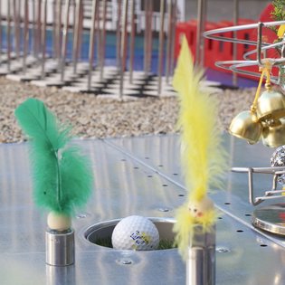 Minigolfball in Loch auf Metallplatte mit zwei Metallstiften, die grüne und gelbe Federn tragen