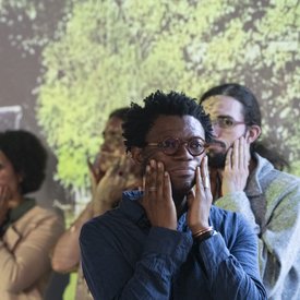 Im Vordergrund Neo Muyanga, der mit seinen Händen sein Gesicht massiert – als Aufwärmübung zum Singen. Im Hintergrund Personen des Projektchors, die ebenfalls Ihr Gesicht massieren.