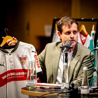 Ingmar Volkmann in beigem Sakko hält ein weiß-rotes Fußballtrikot mit dem VfB Stuttgart-Logo und dem Schriftzug 'WIR STINKLUSION chancengleichheit'.