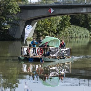 Das Festival-Floß unterwegs auf einem Gewässer in der Region. An Bord sind verschiedene Personen. Eine Person schwingt eine Fahne. Ein Banner an der Reling des Floßes zeigt das Logo von JETZT!.