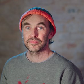 Ein Mann mittleren Alters mit hellem Teint, blauen Augen und kurzem braunem Haar mit etwas Grau blickt direkt in die Kamera. Er trägt eine bunte Strickmütze in Orange und Blau sowie ein graues Sweatshirt. Sein Gesicht ist leicht bärtig. Der Hintergrund ist unscharf und in warmen Tönen gehalten, vermutlich eine Backsteinwand.