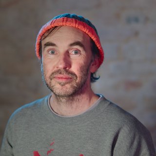 Ein Mann mittleren Alters mit hellem Teint, blauen Augen und kurzem braunem Haar mit etwas Grau blickt direkt in die Kamera. Er trägt eine bunte Strickmütze in Orange und Blau sowie ein graues Sweatshirt. Sein Gesicht ist leicht bärtig. Der Hintergrund ist unscharf und in warmen Tönen gehalten, vermutlich eine Backsteinwand.