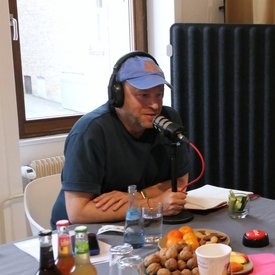 Jacob Bilabel, Person mit blauer Kappe und Kopfhörern sitzt an Tisch mit Mikrofon, Notizbuch, Getränken, Nüssen und Obstschale.