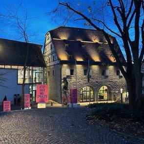 Abendaufnahme eines historischen Gebäudes in Böblingen mit beleuchtetem Plakat, das zur Ausstellung ‚Schlamassel Tov‘ einlädt. Verschiedene pinkfarbene Plakate sind vor dem Gebäude sichtbar.